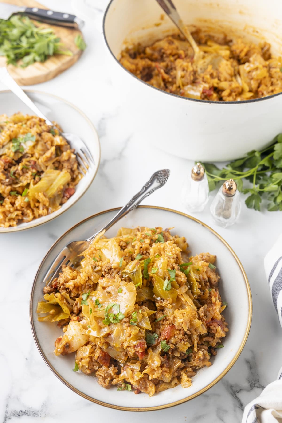 Dutch oven filled with deconstructed cabbage rolls beside bowls of the dish and fresh parsley.