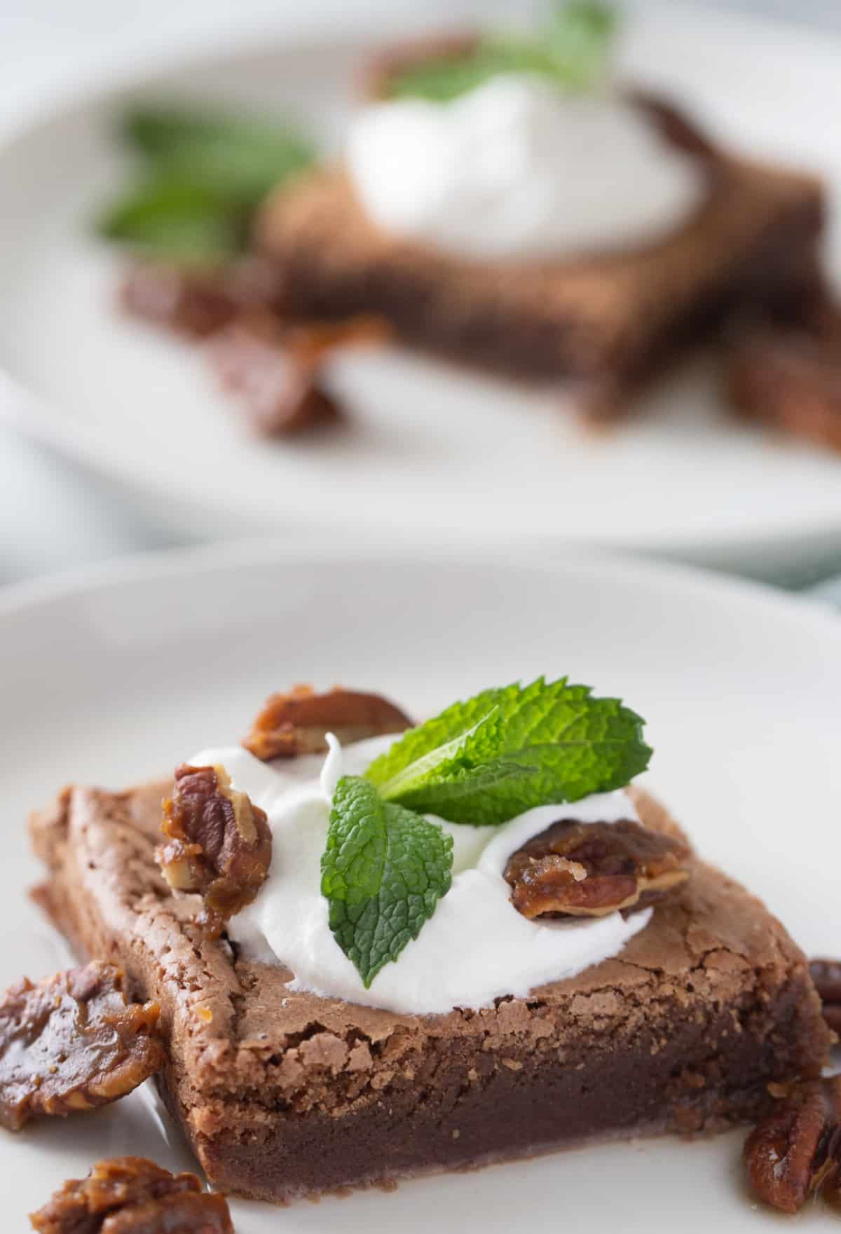 Mint Julep Brownie with whipped cream, pecans, and fresh mint Brownie on a plate topped with whipped cream, candied pecans, and mint, with another dressed brownie blurred in the back