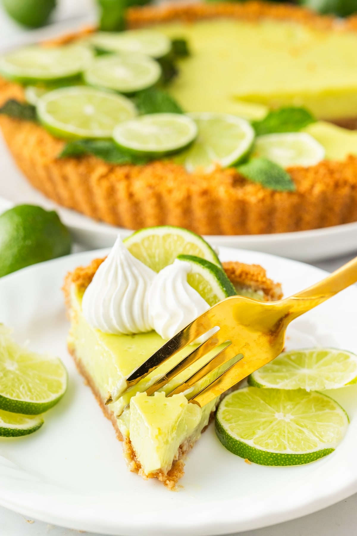 Gold fork cutting into a slice of Key lime tart with the full tart in the background