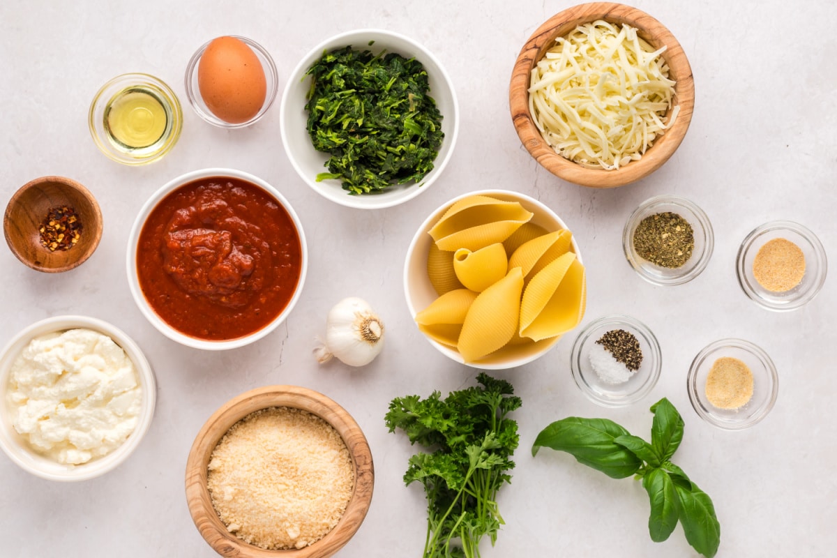 Ingredients for stuffed shells with ricotta, spinach, mozzarella, Parmesan and marinara arranged on a white surface.
