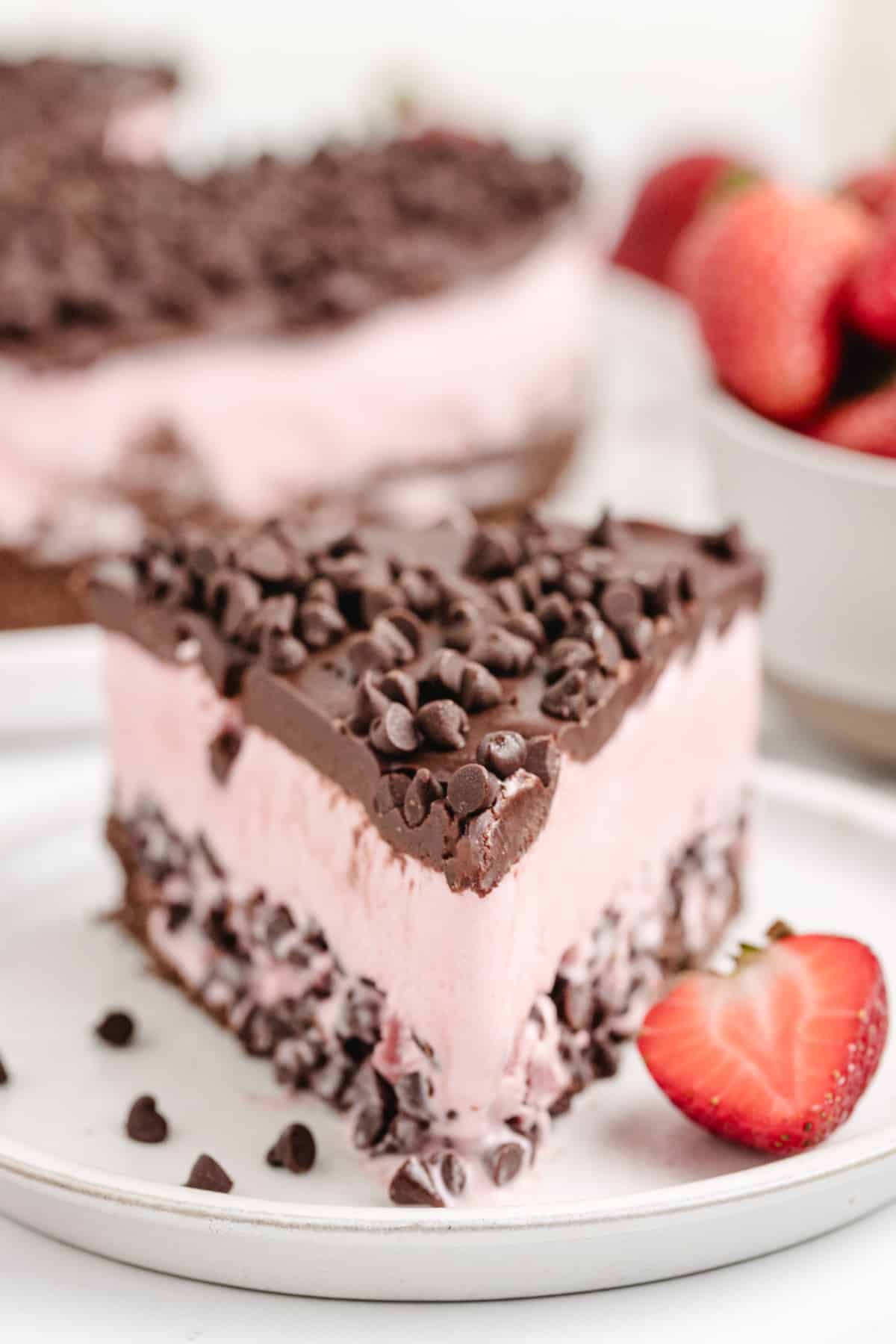 A creamy and cold slice of strawberry chocolate chip ice cream cake on a plate, showcasing its layers of chocolate graham cracker crust, strawberry ice cream with chocolate chips, and rich chocolate topping.