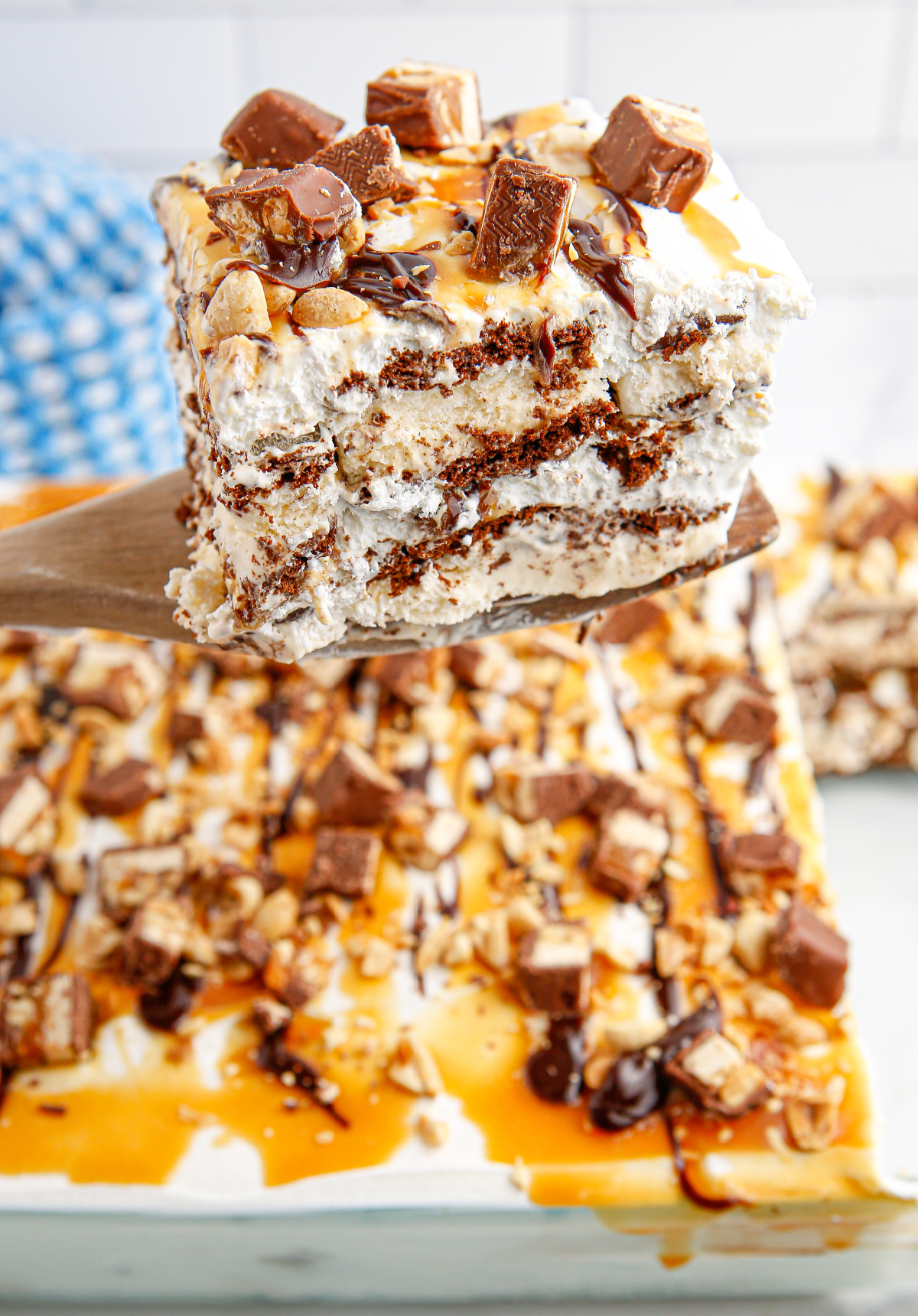 Large square of snickers ice cream cake on spatula above a full pan topped with candy and sauces