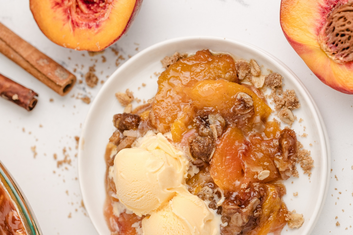 Peach dessert with oat topping and ice cream. Bowl of warm peach crisp topped with vanilla ice cream, surrounded by fresh peaches and cinnamon sticks.