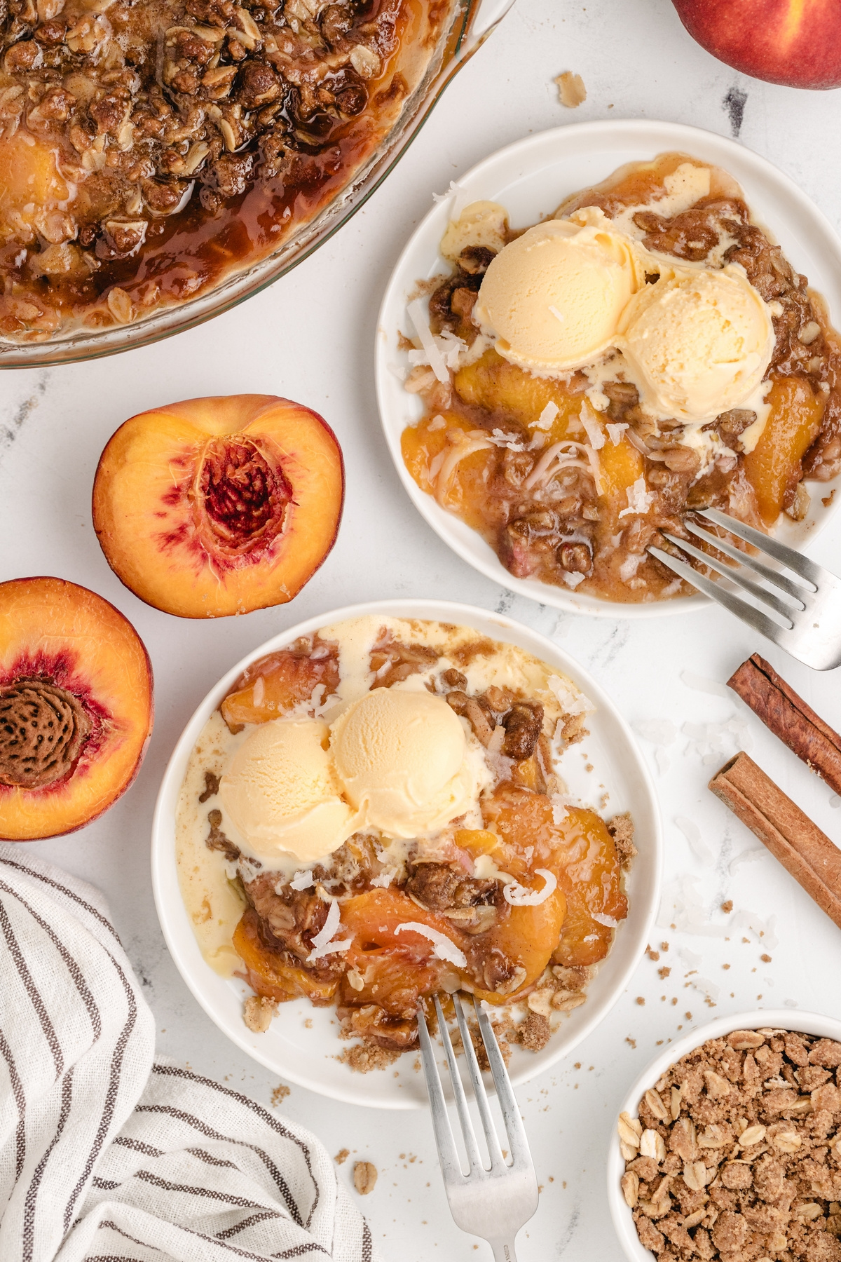 Plates of peach crisp with oat topping and ice cream. Two plates of peach crisp with ice cream, fresh peaches, and cinnamon sticks on table.