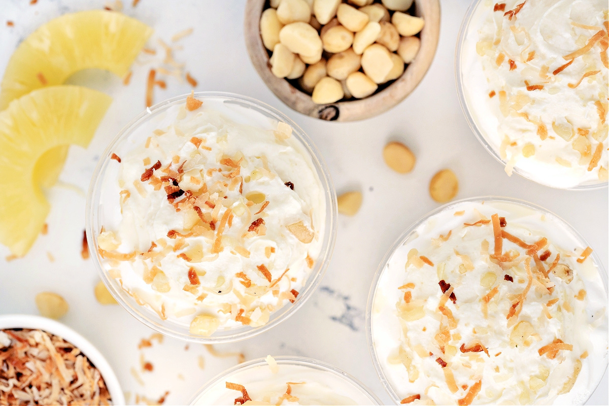 Overhead view of Hawaiian Dream Dessert Cups with toasted coconut and macadamias Overhead view of Hawaiian Dream Dessert Cups topped with whipped cream, toasted coconut, and macadamia nuts surrounded by pineapple slices.
