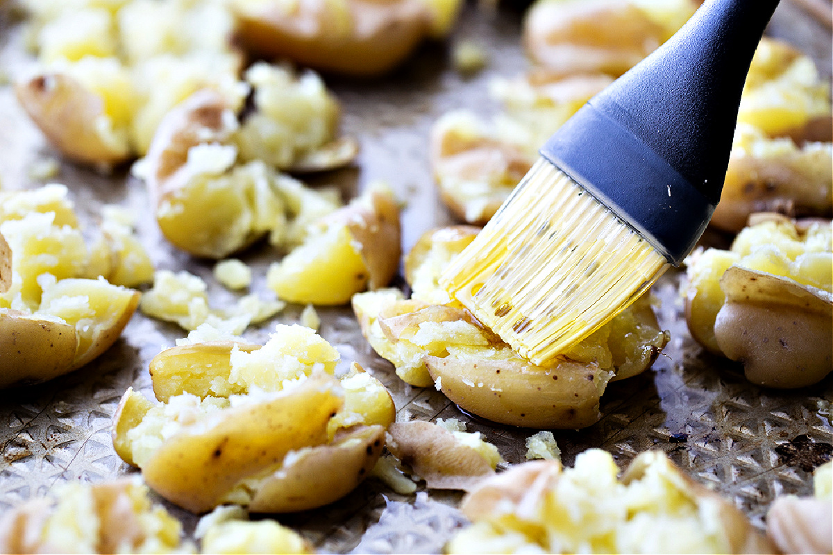 brushing vinegar on potatoes, finishing potatoes, vinegar step, sheet pan potatoes A pastry brush coating hot smashed potatoes with apple cider vinegar on a sheet pan, the final step that gives salt and vinegar smashed potatoes their tang.