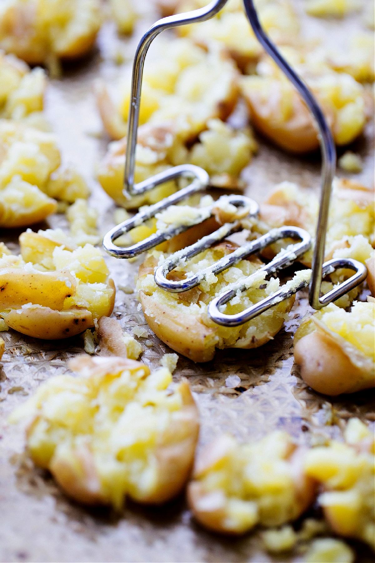potato masher, smashed potatoes, sheet pan prep, rustic potatoes A potato masher pressing boiled potatoes on a sheet pan to make salt and vinegar smashed potatoes, showing the rough, fluffy texture before roasting.