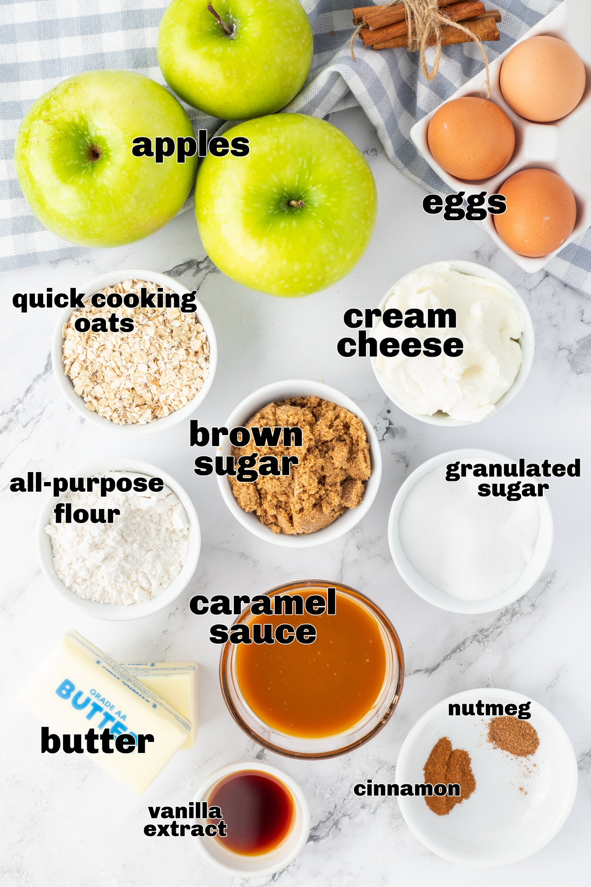 Ingredients for apple cheesecake bars laid out on a marble counter, including apples, cream cheese, butter, oats, sugar, and caramel sauce.