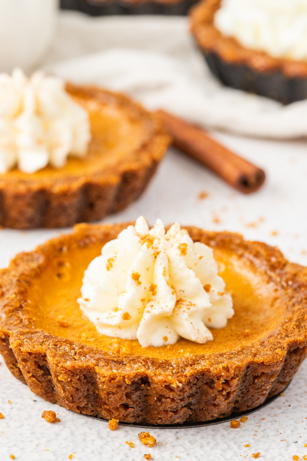Close-up of a pumpkin tart with gingersnap crust and whipped cream topping, sprinkled with cookie crumbs and cinnamon.