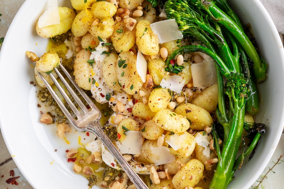 Bowl of potato gnocchi with burrata and pesto ready to eat. Close-up of crispy gnocchi with burrata, broccolini, toasted nuts, and pesto in a white bowl with a fork.