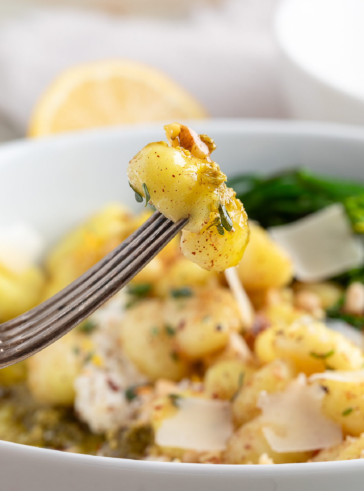 Fork lifting brown butter gnocchi with burrata and pesto. Fork holding a bite of gnocchi coated in brown butter with fresh herbs, over a bowl with burrata, pesto, shaved parmesan, and broccolini.
