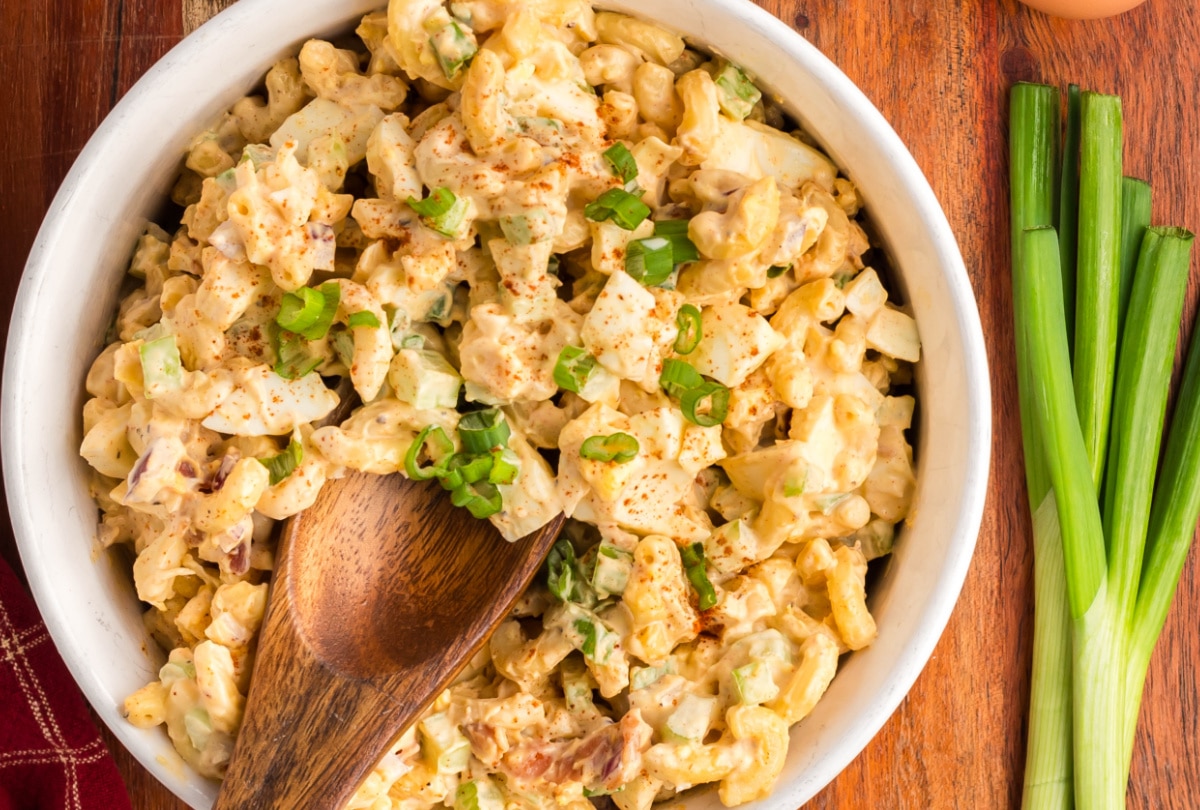 A bowl of deviled egg pasta salad with a wooden spoon resting on the side and fresh green onions beside it.