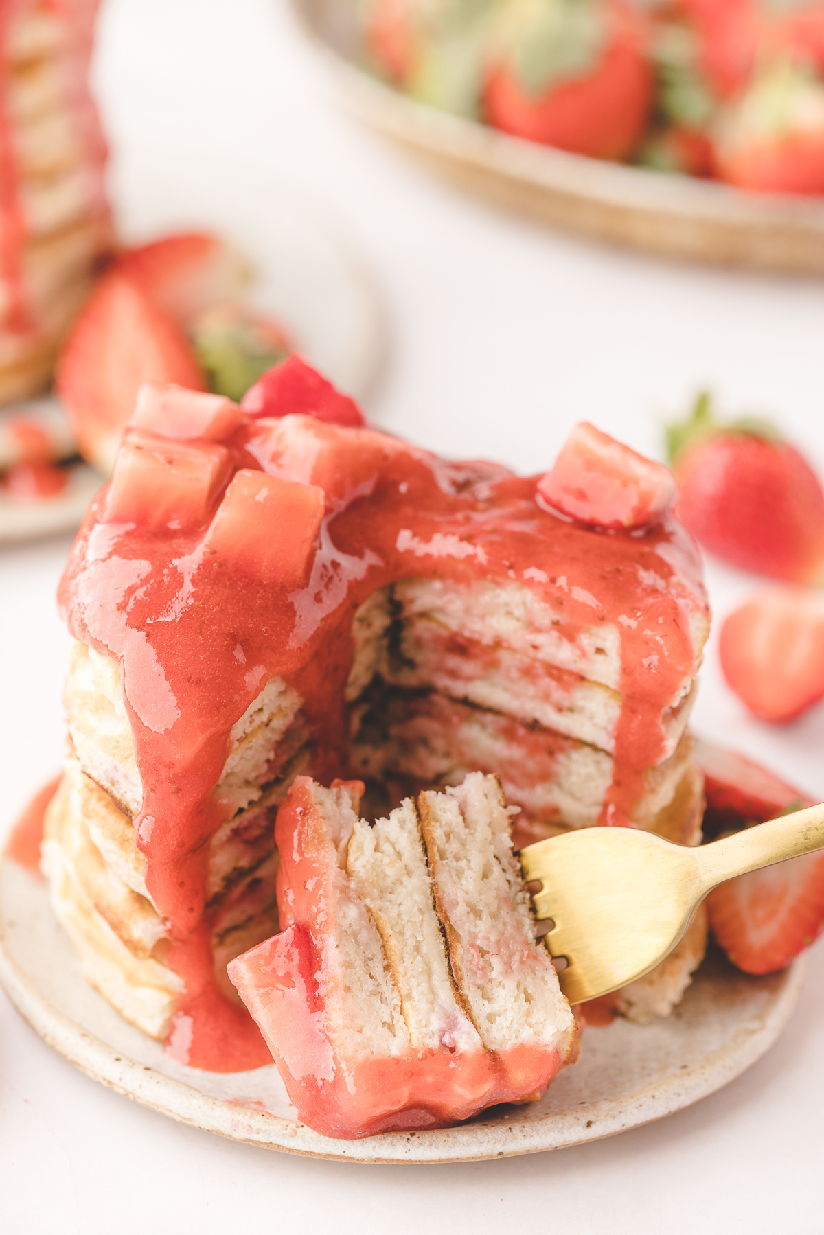 Strawberry Pancakes with Jammy Strawberries Fork lifting a bite of strawberry pancakes with jammy strawberries inside the buttermilk stack and warm strawberry sauce dripping over the top.