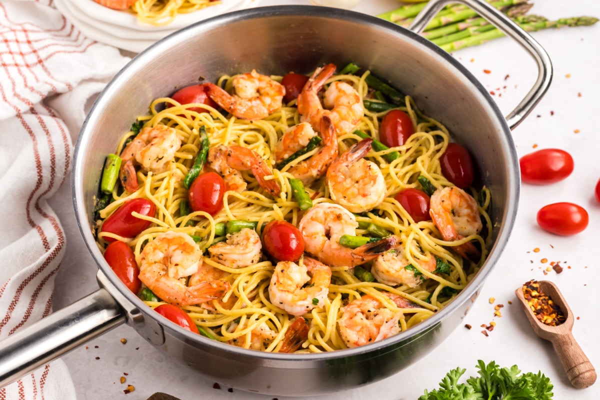 ready to eat Garlic Shrimp Pasta with Asparagus and Tomatoes Side angle of garlic shrimp pasta in a skillet, surrounded by red pepper flakes and asparagus on the counter.