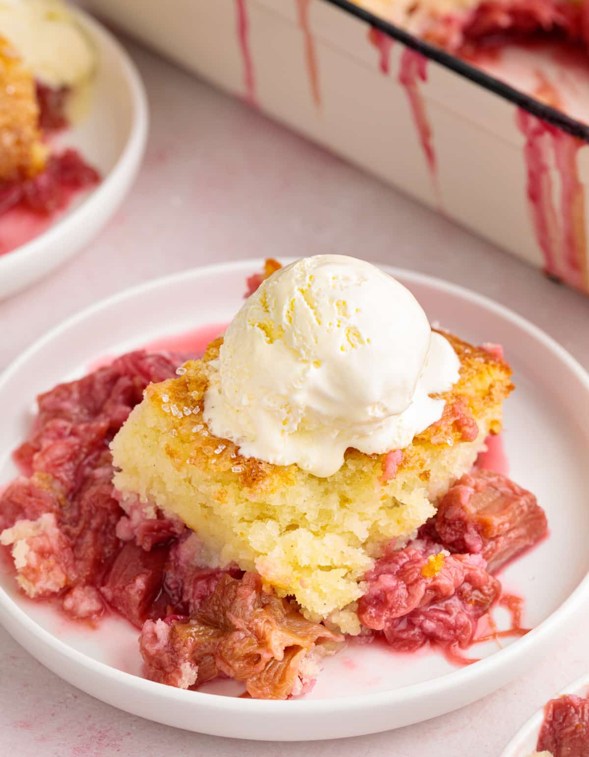 A close-up of a spoonful of rhubarb cobbler with ice cream, with the baking dish slightly blurred in the background.