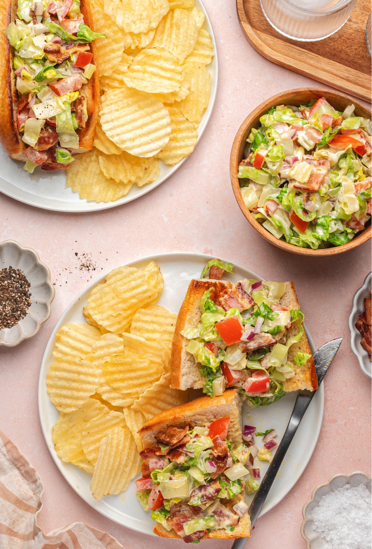 Flat lay of BLT hoagies with chips and ingredients Flat lay of chopped BLT hoagie sandwiches on plates, cut in half with potato chips, water, and ingredients scattered around