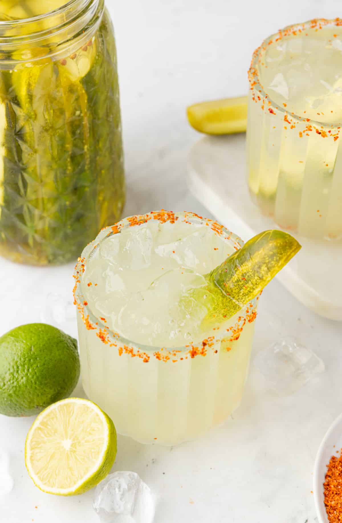 Two dill pickle margaritas with a pickle jar, lime, and ice on the table