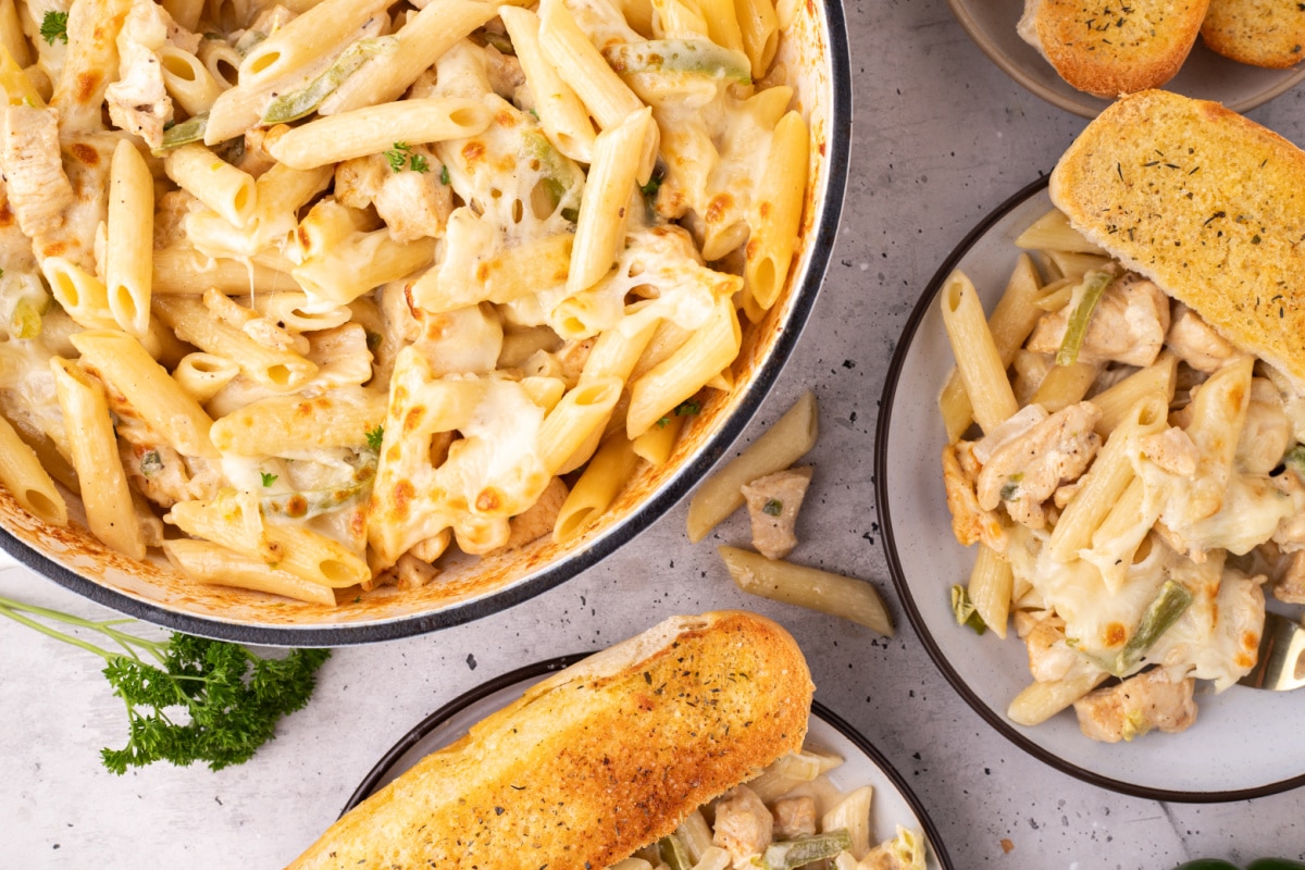 Skillet of chicken Philly cheesesteak pasta with plated portions and garlic bread Overhead view of skillet filled with chicken Philly cheesesteak pasta surrounded by plated servings and garlic bread