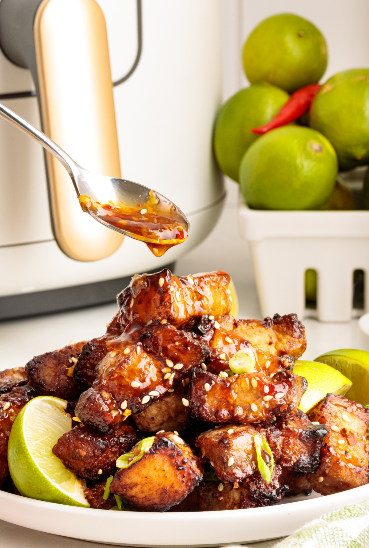 Sticky glazed pork belly bites being drizzled with sweet chili lime sauce in front of an air fryer, surrounded by fresh limes and chili peppers.