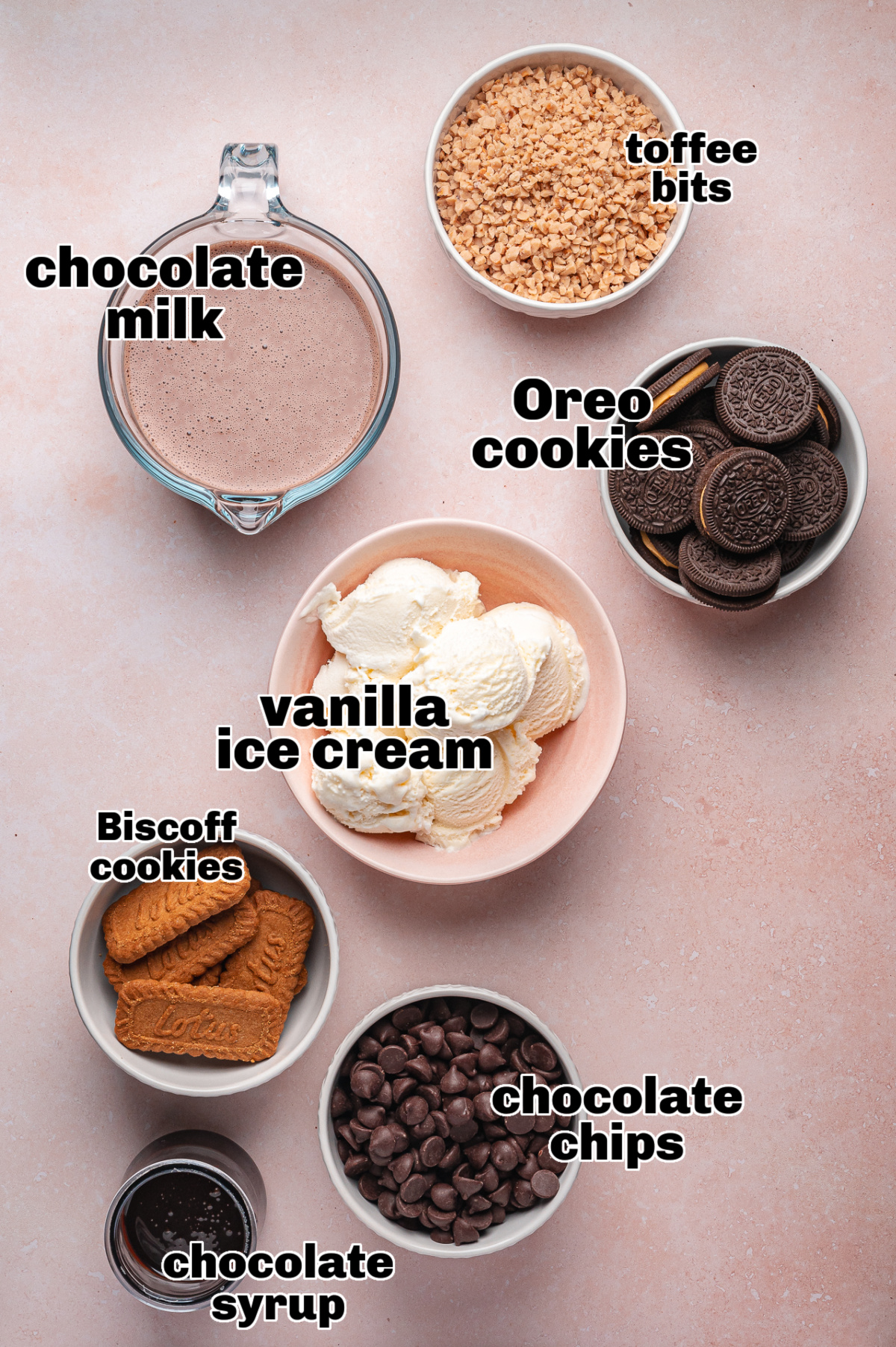 Chocolate freakshake ingredients laid out on pink background Bowls of chocolate freakshake ingredients including ice cream, Oreos, Biscoff cookies, toffee bits, and chocolate milk