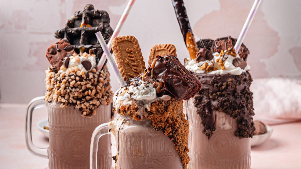 Chocolate milkshake mugs with different cookie rims and loaded toppings Multiple chocolate freakshakes in glass mugs with candy, cookies, and brownies