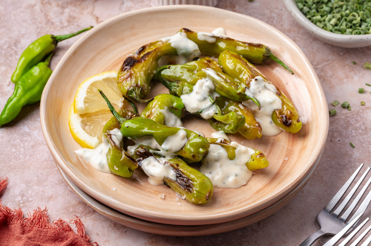 Blistered shishito peppers served with garlic chive aioli and lemon slices on a wooden plate