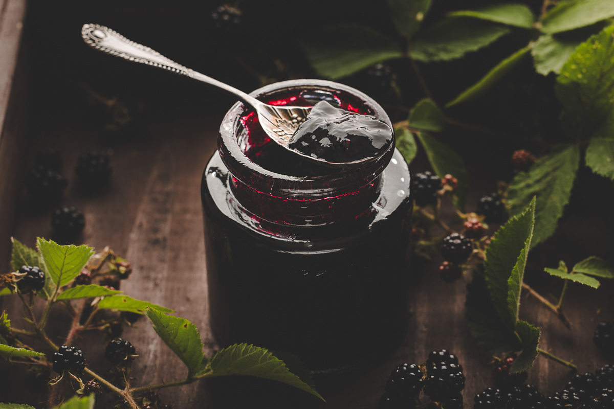 Dark moody jar of blackberry jam with spoonful lifted, framed by wild blackberry vines