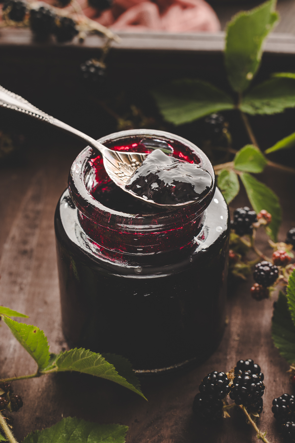 Spoon lifting glossy blackberry jam from a jar surrounded by fresh blackberry leaves