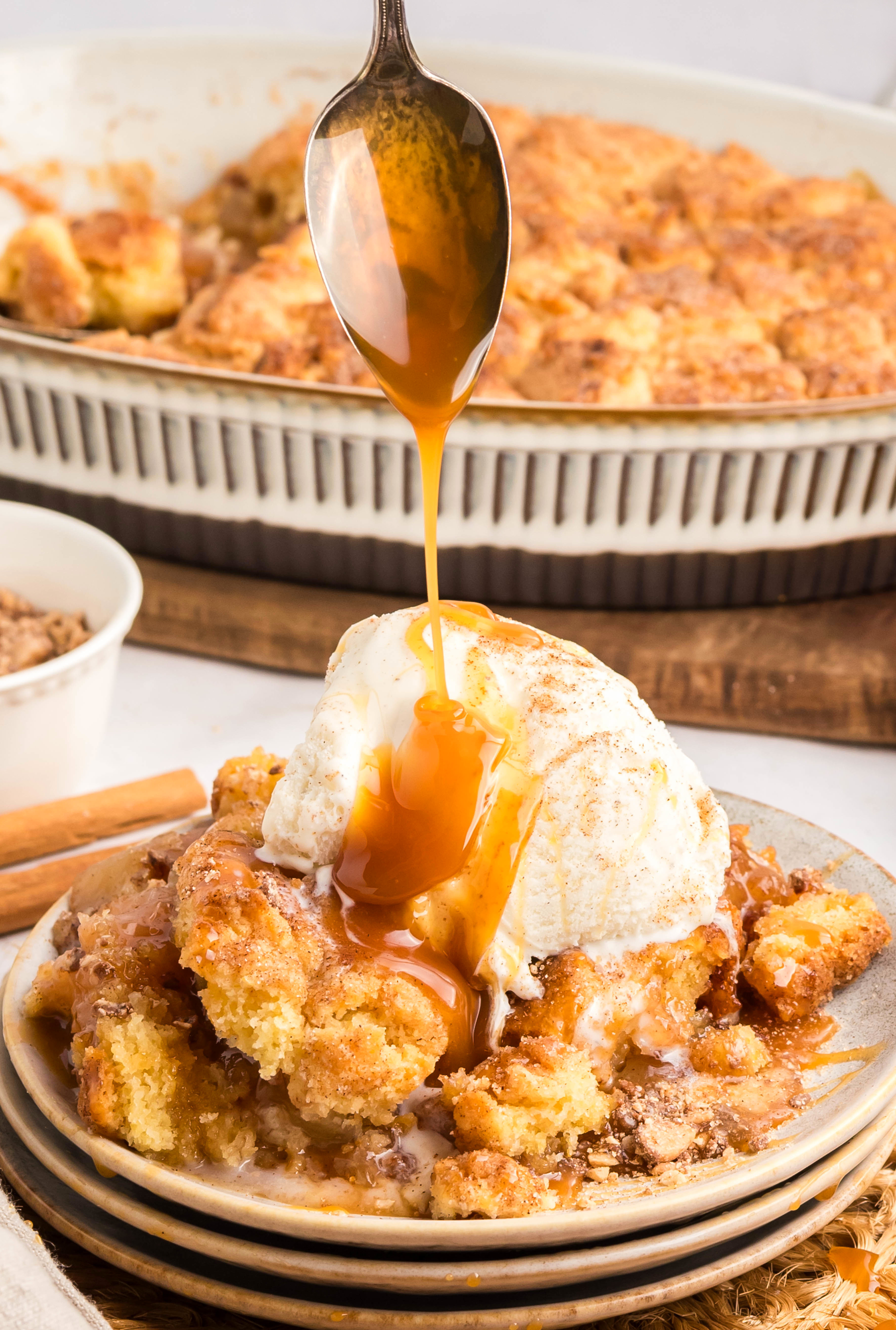 Spoon drizzling caramel over warm snickerdoodle apple cobbler with ice cream.