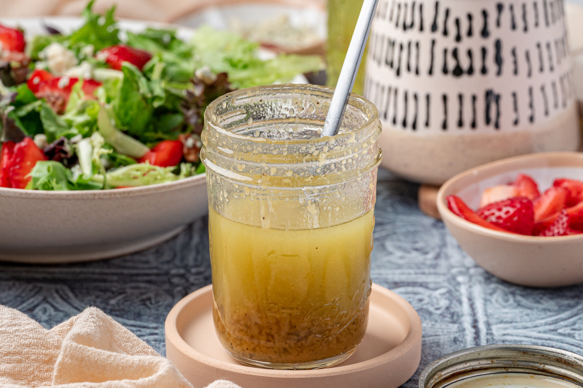 Homemade champagne vinaigrette in a jar with spoon, surrounded by fresh salad greens and sliced strawberries.