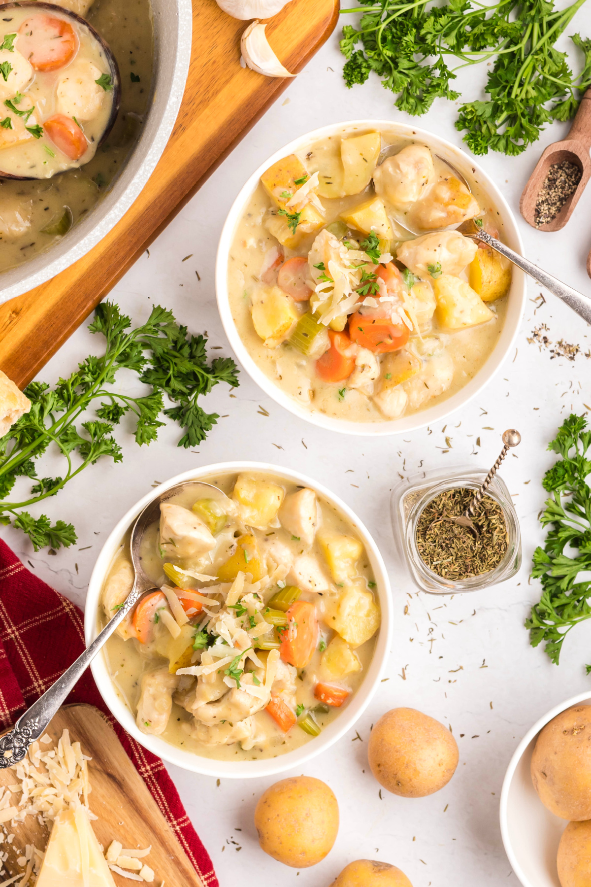 bowl of chicken and vegetable stew with potatoes, carrots, celery, and Parmesan, surrounded by fresh parsley, garlic, and herbs on the counter.