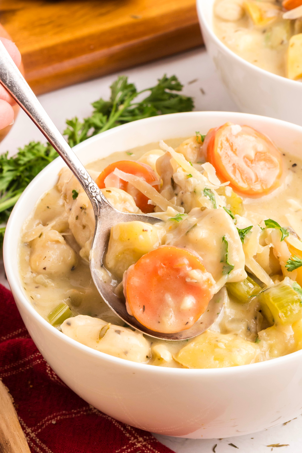 spoon lifting creamy chicken and vegetable stew with carrots, celery, potatoes, and Parmesan from a white bowl, showing thick broth and herbs.