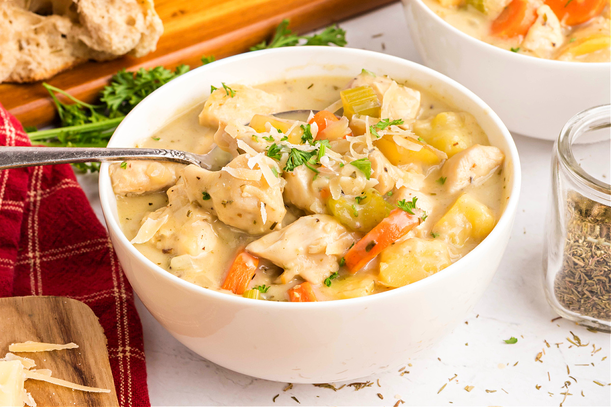 close-up of creamy chicken stew in a white bowl with chicken, carrots, celery, and potatoes in a thick, creamy broth topped with parsley and Parmesan.
