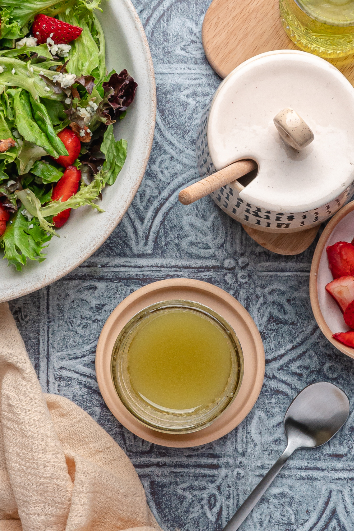 Glass jar of Dijon champagne dressing next to a strawberry and mixed greens salad on a textured blue surface.