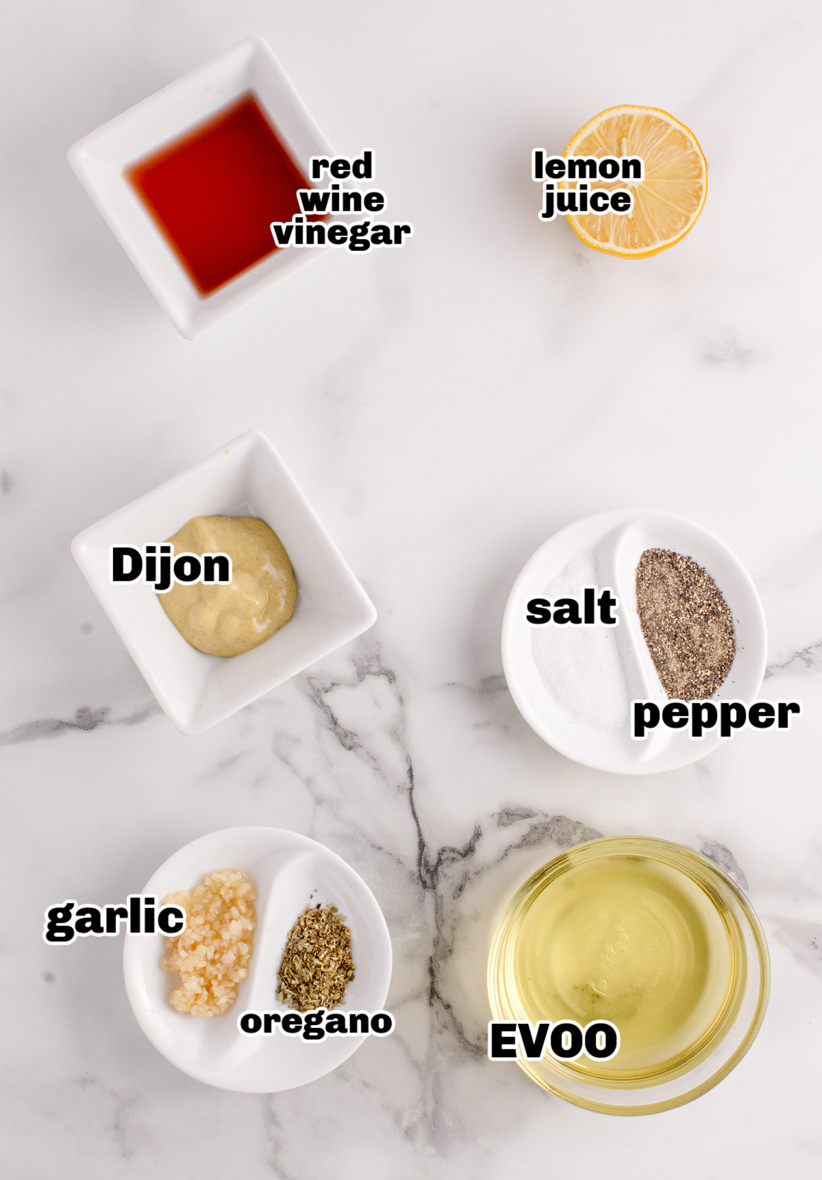 Ingredients for Greek salad dressing recipe on a marble surface, including olive oil, red wine vinegar, lemon, Dijon mustard, garlic, oregano, salt, and pepper.