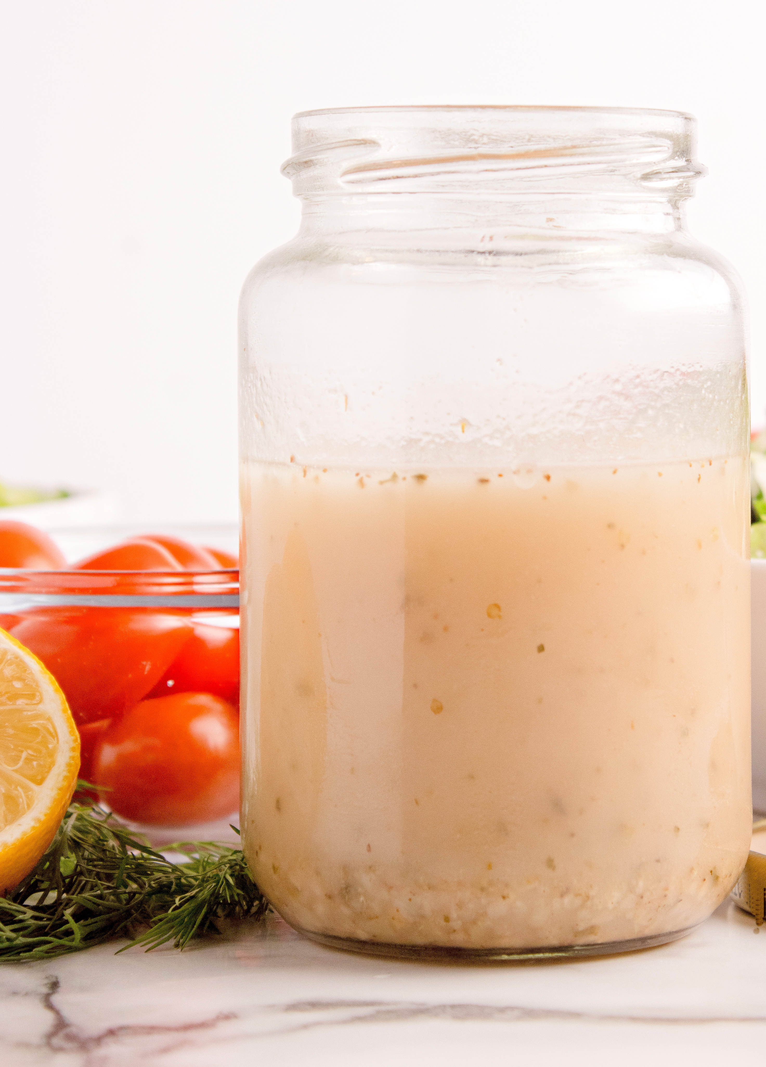 Side view of a glass jar filled with creamy Greek salad dressing and tomatoes and lemon in the background.