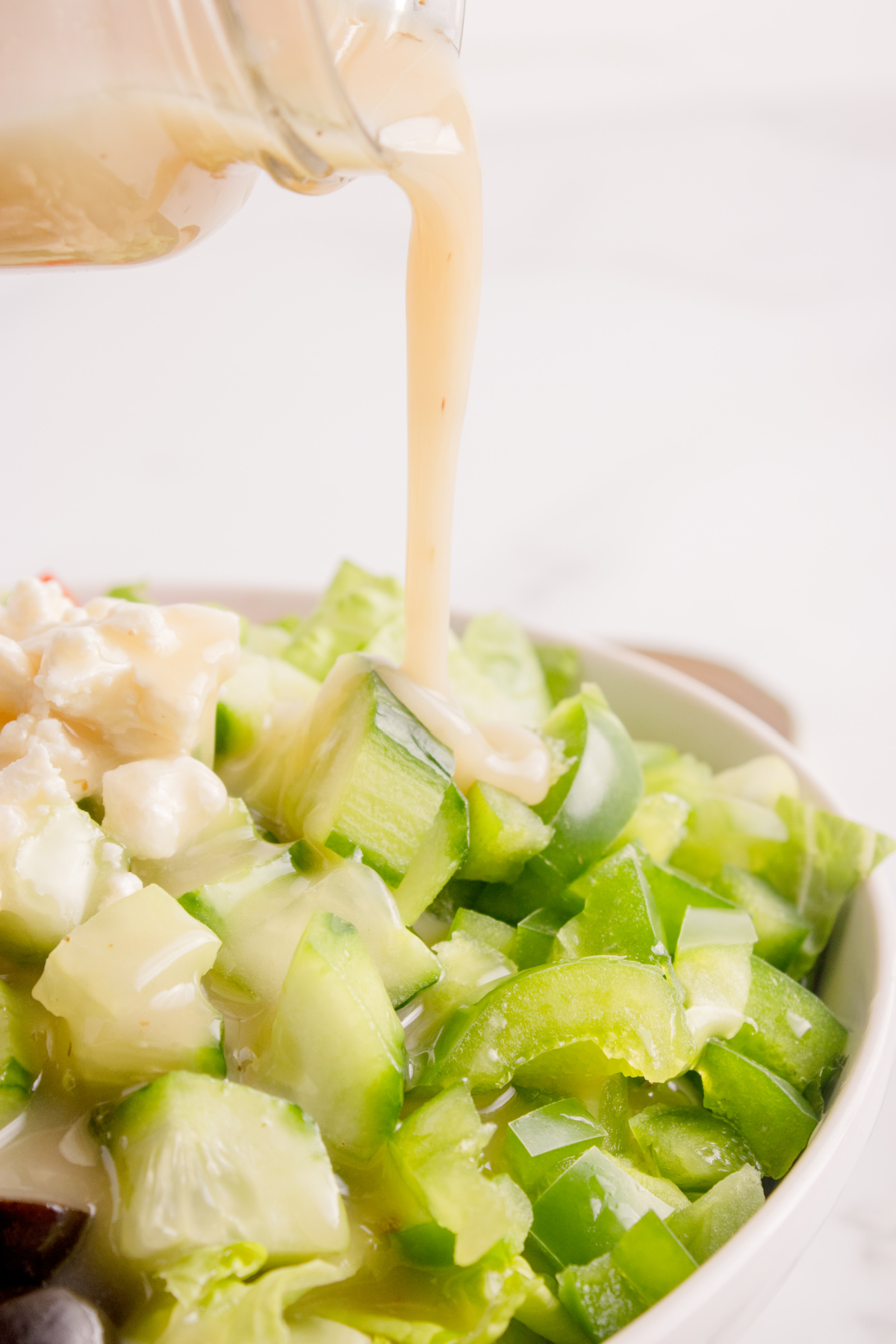Greek salad dressing being poured from a jar over a bowl of chopped cucumbers, peppers, and feta.