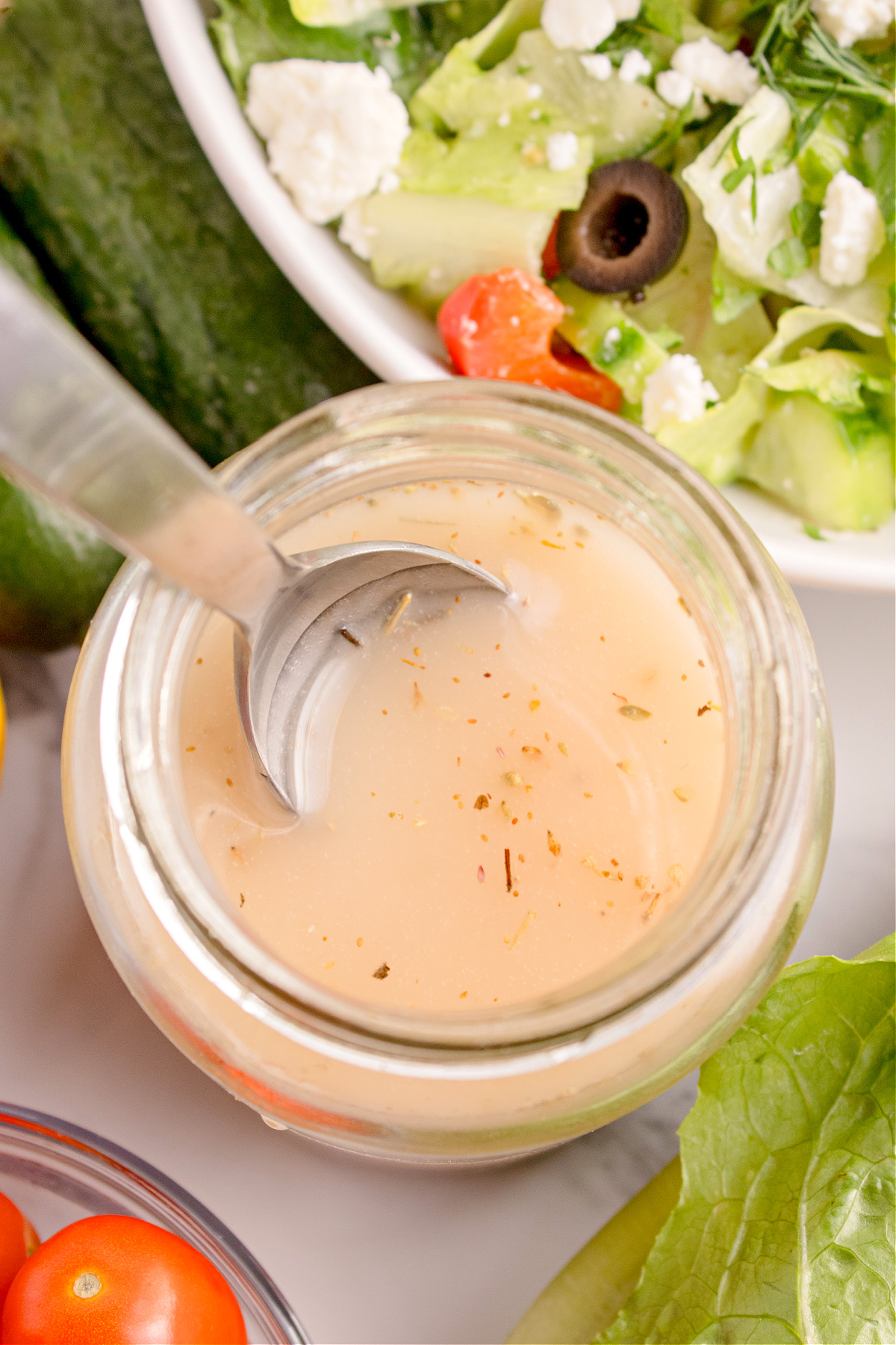 Jar of homemade Greek vinaigrette with a spoon beside a Greek salad topped with feta, olives, and tomatoes.