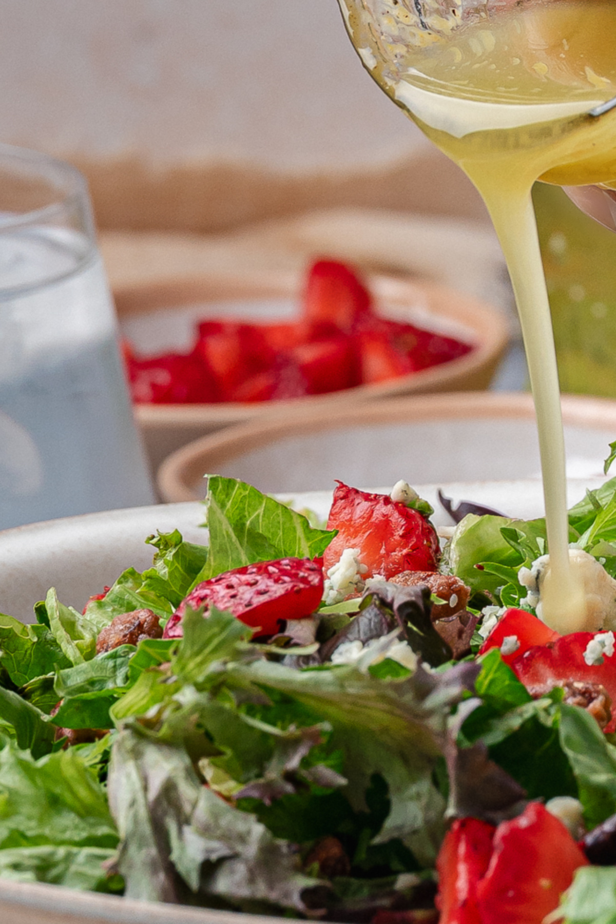 Pouring homemade salad dressing over a bowl of mixed greens with strawberries and cheese.