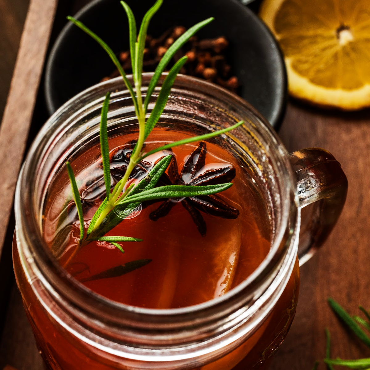 Glass mug of mulled cider garnished with rosemary and star anise beside sliced lemon and cloves, photographed for Noble Pig.