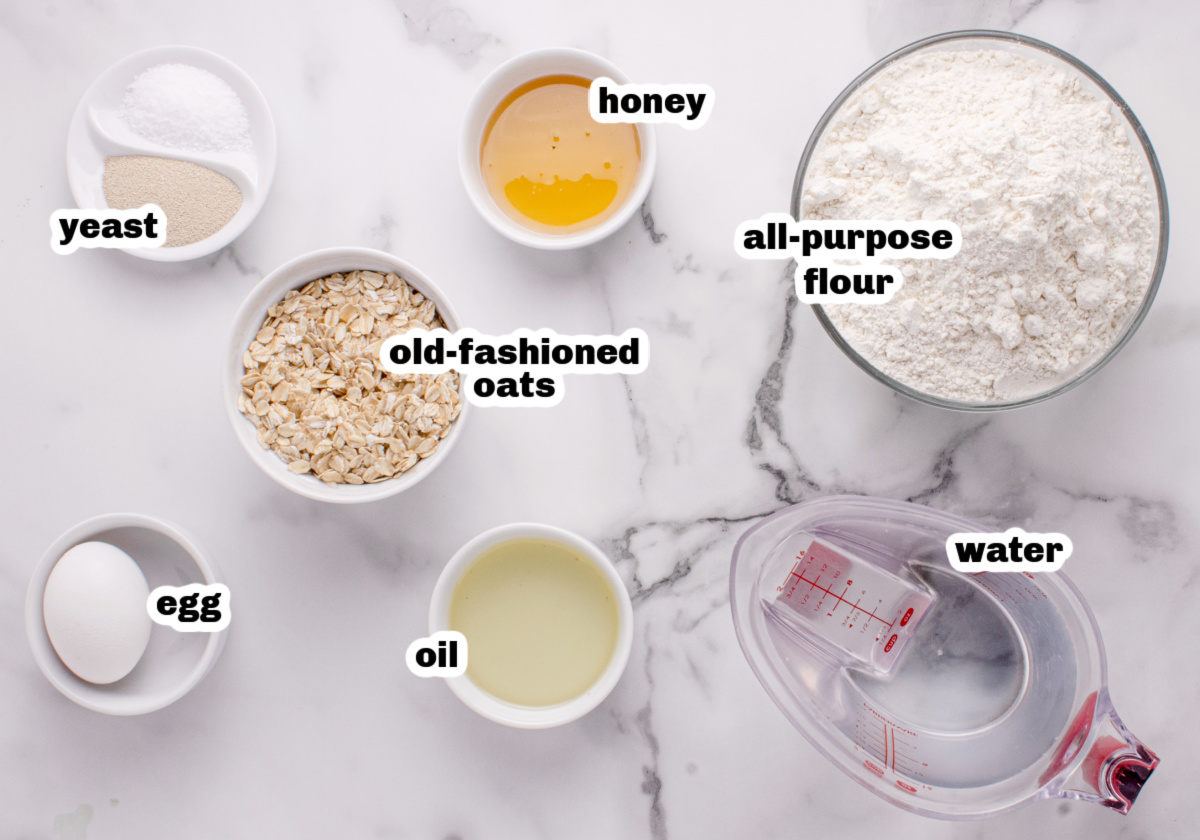 honey oatmeal bread ingredients Ingredients for homemade oatmeal bread including oats, flour, honey, yeast, oil, and egg on a marble counter.