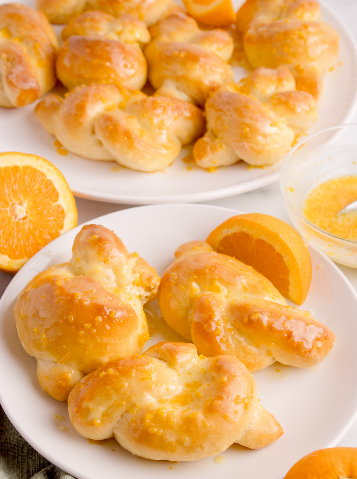 Orange Knot Rolls with Citrus Glaze Orange knot rolls on a white plate, brushed with orange glaze and surrounded by orange slices.