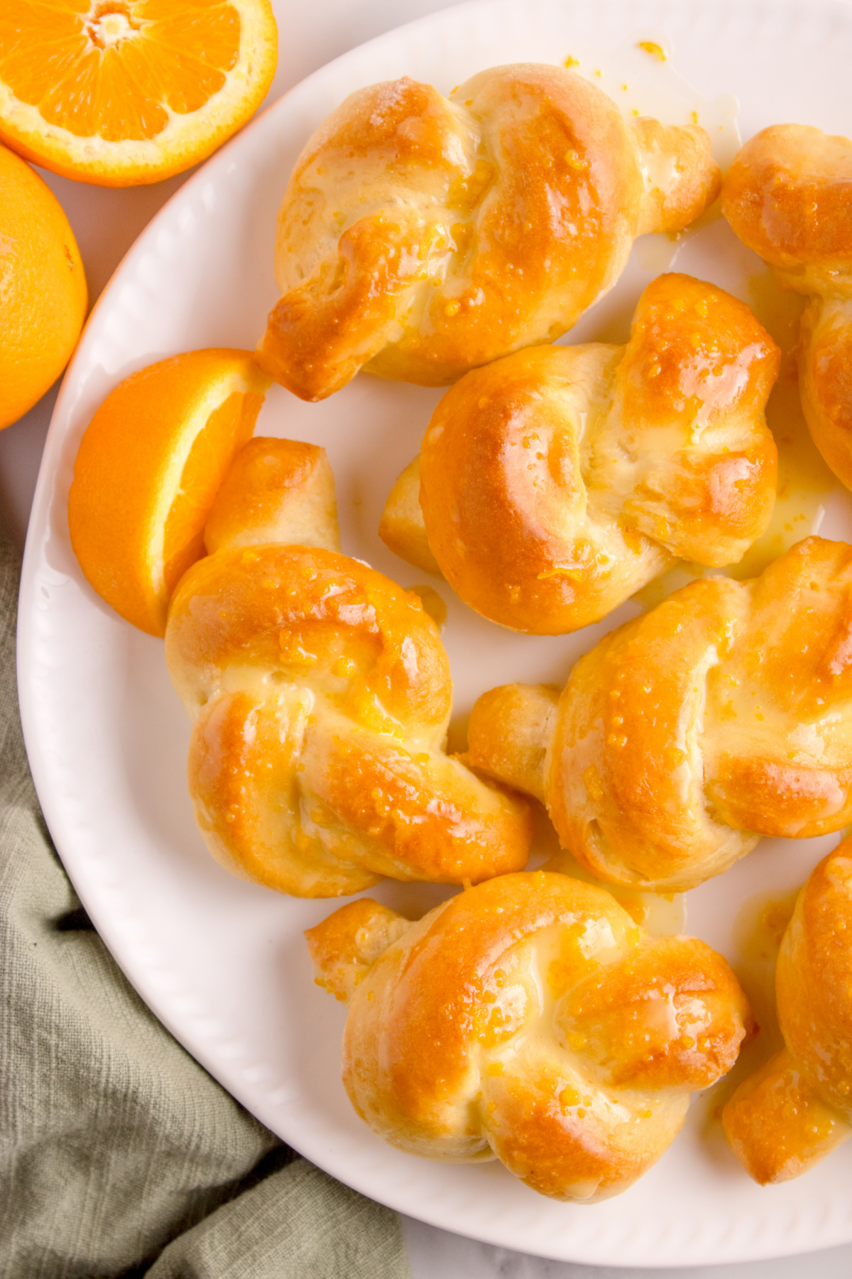 Glazed Orange Knot Rolls Plate of golden orange knot rolls with shiny glaze and sliced oranges beside them.