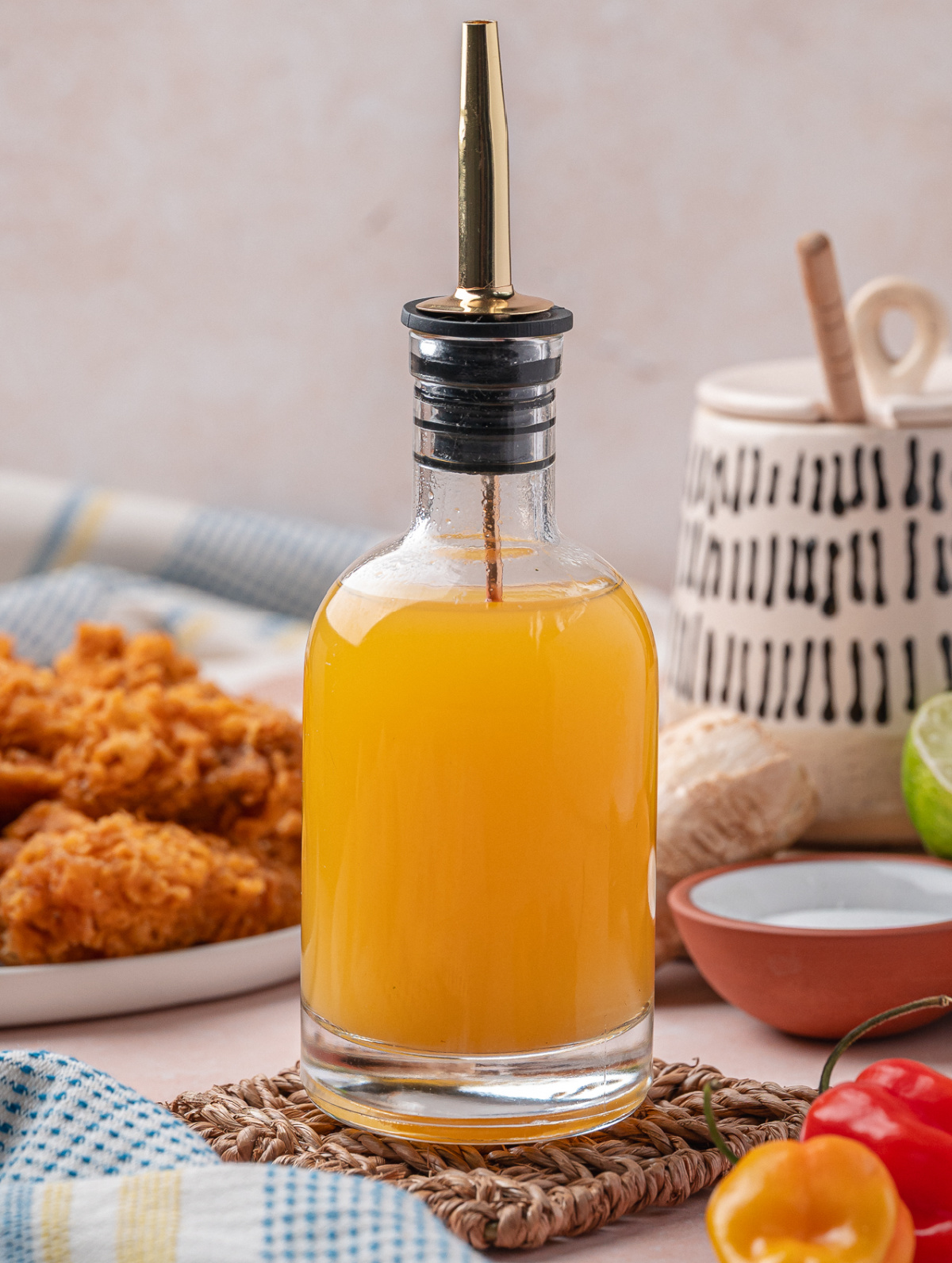 Clear glass bottle filled with golden homemade hot sauce, with fried chicken and habanero peppers in the background.
