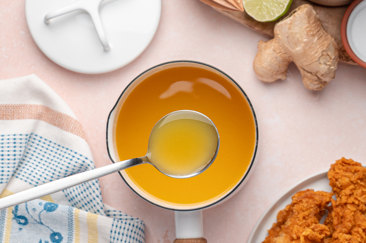 Ladle lifting smooth pineapple habanero hot sauce from a white saucepan beside ginger and lime.