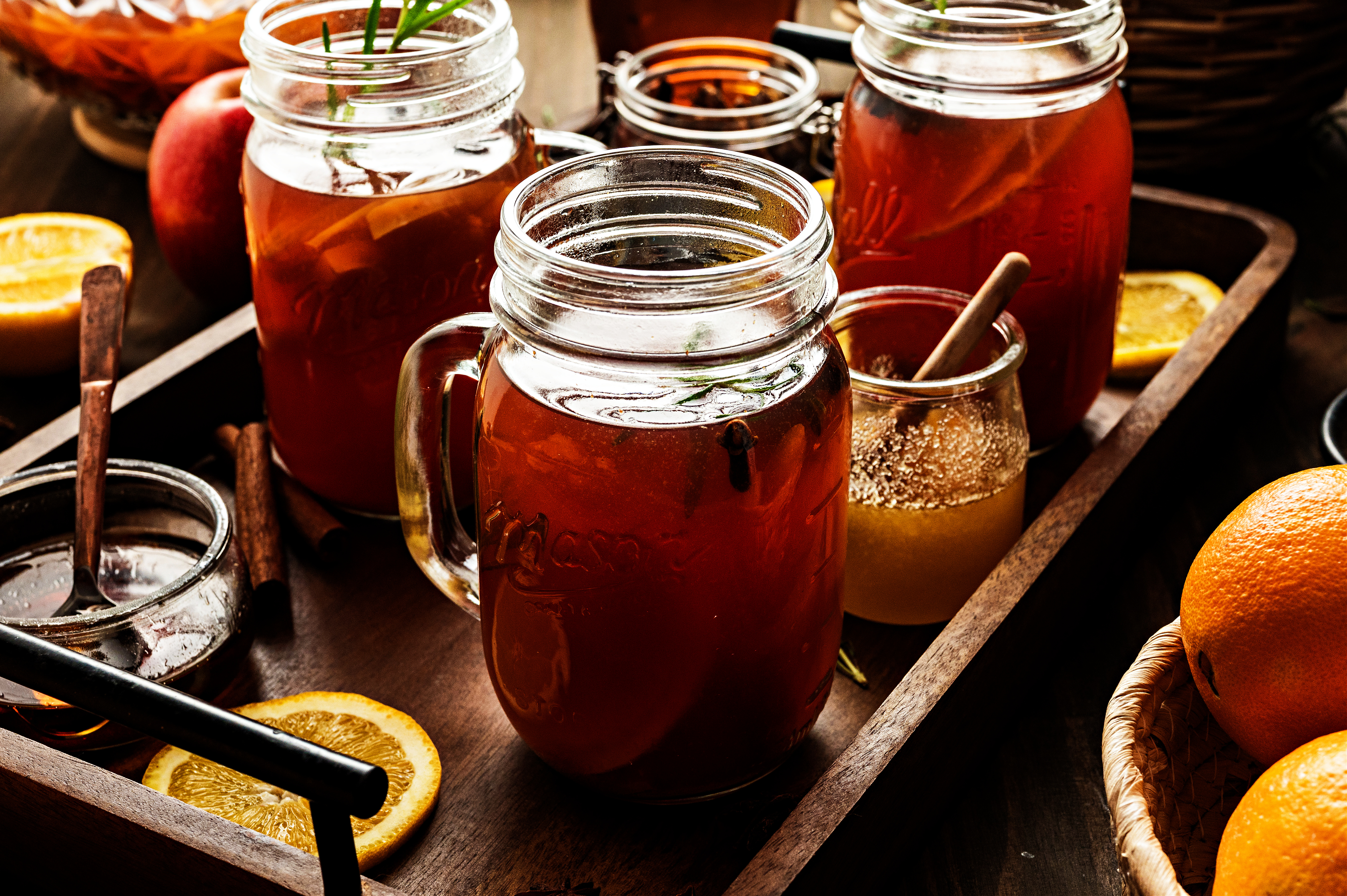 Glass mugs filled with mulled cider beside oranges, honey, and cinnamon sticks on a rustic wooden tray. Photographed for Noble Pig.