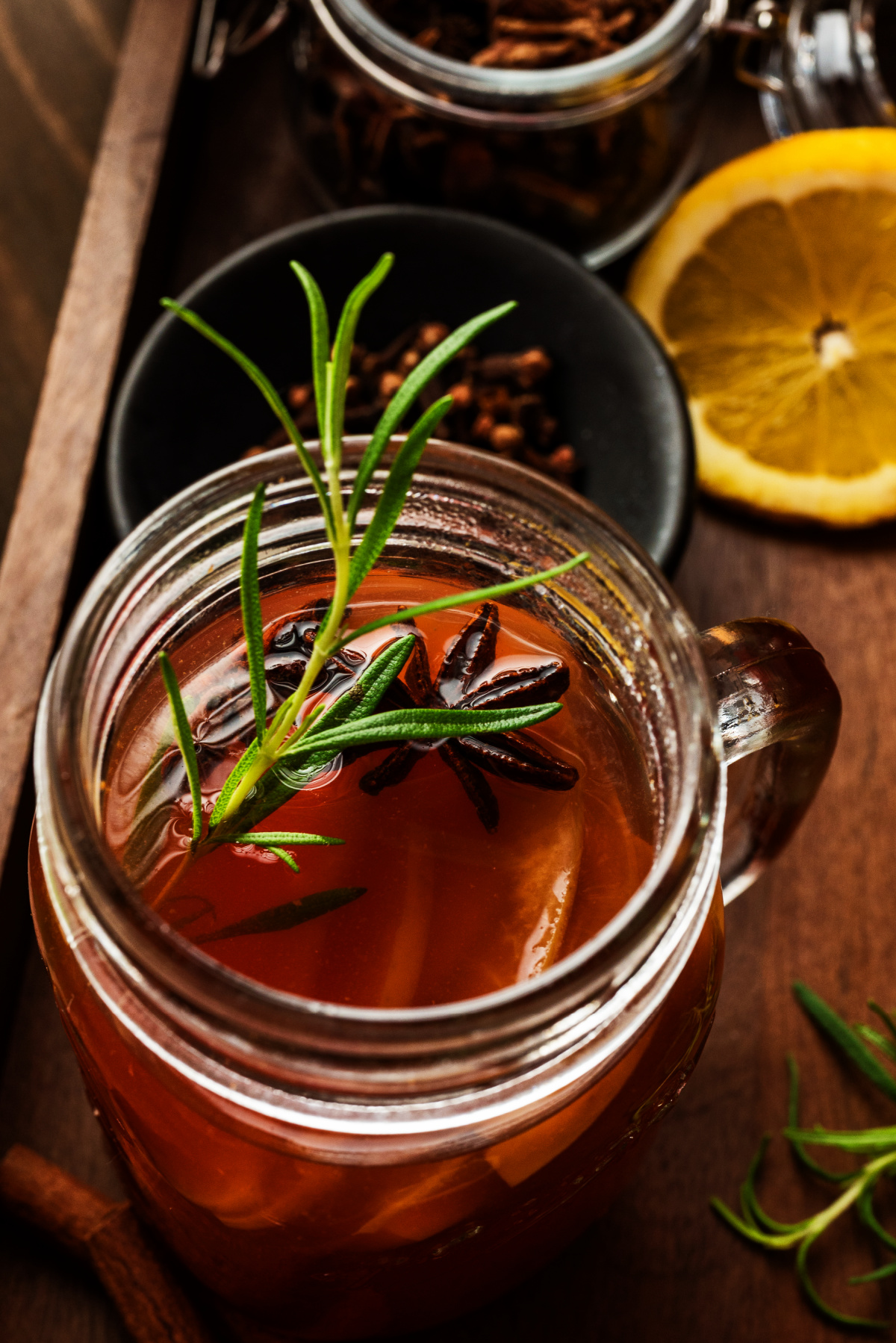 Glass mug of mulled apple cider topped with rosemary and star anise beside lemon slices and cloves, showing the drink’s deep color and aromatic garnish.