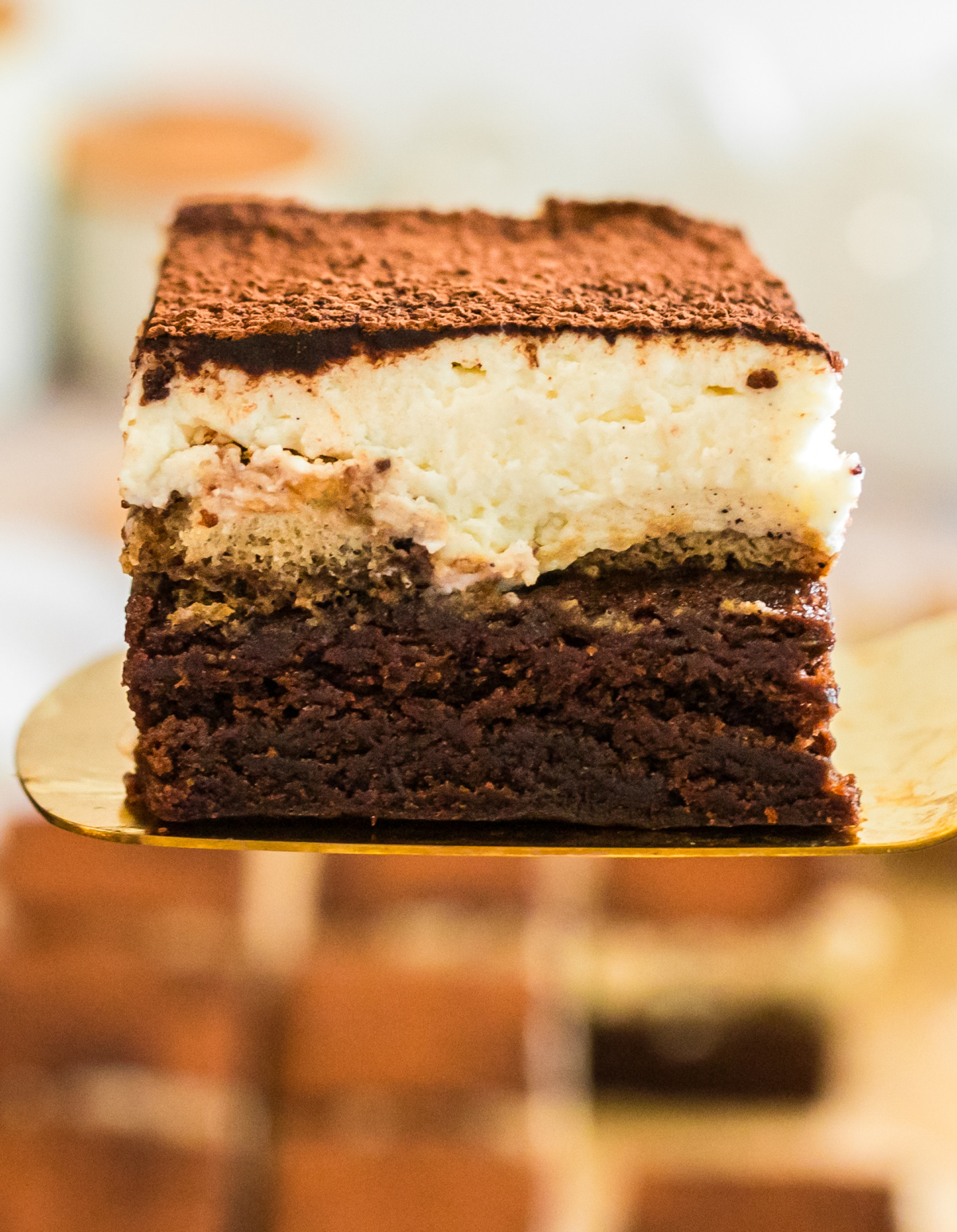 tiramisu brownies slice with cocoa topping and espresso-soaked ladyfingers. slice of tiramisu brownies on a gold spatula with distinct brownie, ladyfinger, and mascarpone layers.