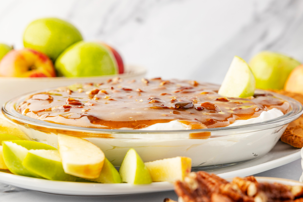 Pecan pie dip in a glass pie dish with a cold cream cheese base topped with warm caramel pecans, surrounded by sliced green apples for dipping by Noble Pig.