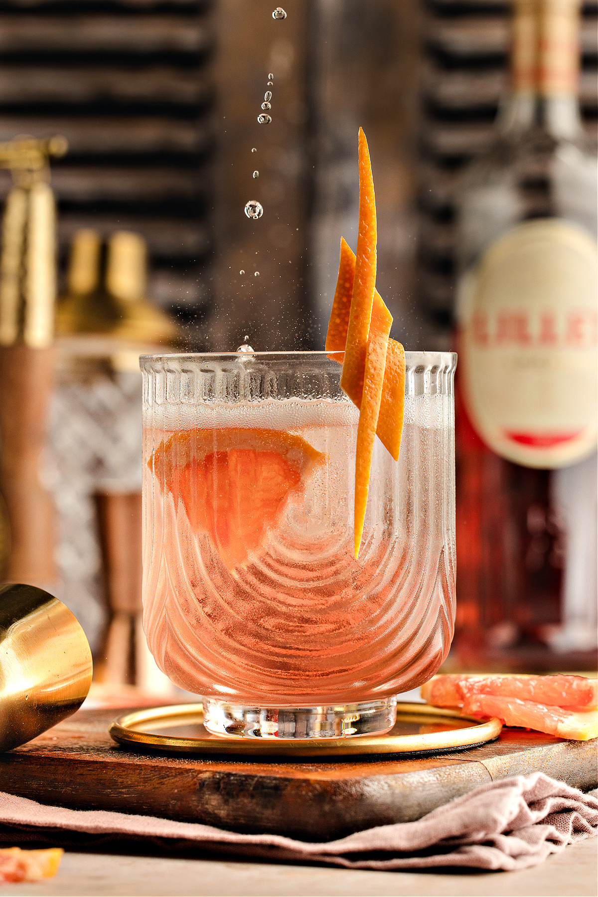 A chilled Lillet Rosé spritz in a textured glass with grapefruit garnish, catching falling droplets as the pale pink drink glows against a blurred bar background.